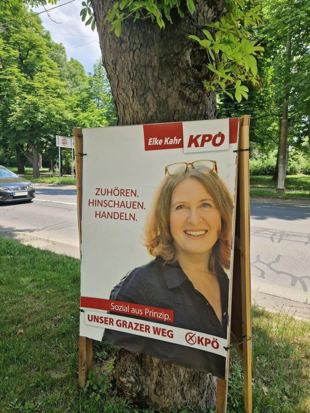 Ein Wahlplakat der KPÖ mit Elke Kahr lehnt an einem Baum am Straßenrand.