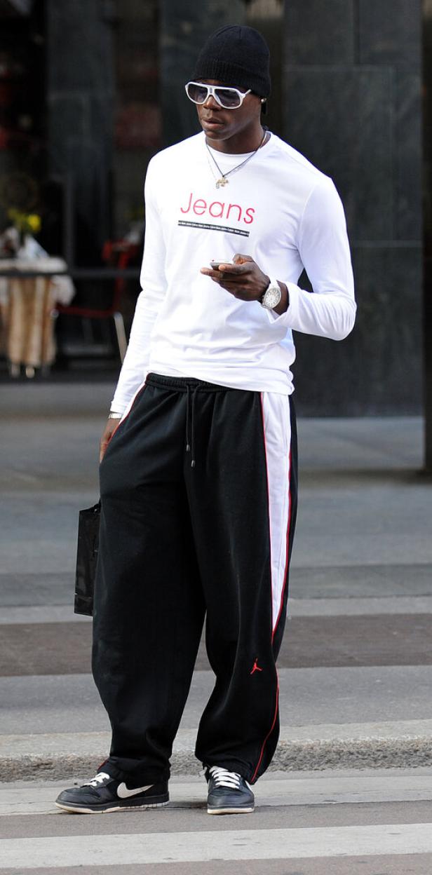 Mario Balotelli mit weißem Langarmshirt, schwarzer Mütze und Sonnenbrille steht auf der Straße und schaut auf sein Handy.