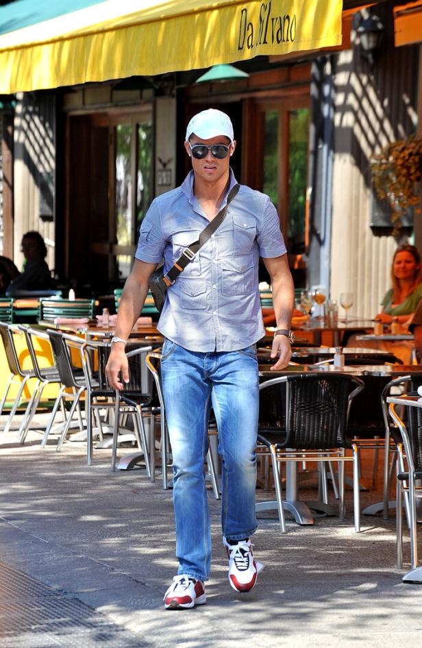 Cristiano Ronaldo mit Sonnenbrille, Basecap und Umhängetasche geht an einem Straßencafé mit leeren und besetzten Tischen vorbei.