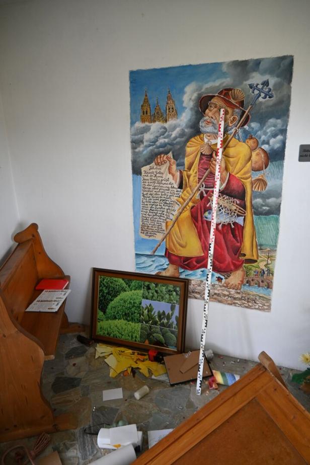Eine bunte Wandmalerei eines Pilgers mit Muschel und Stab, davor Unordnung mit umgestürztem Holzmobiliar und verstreuten Gegenständen.