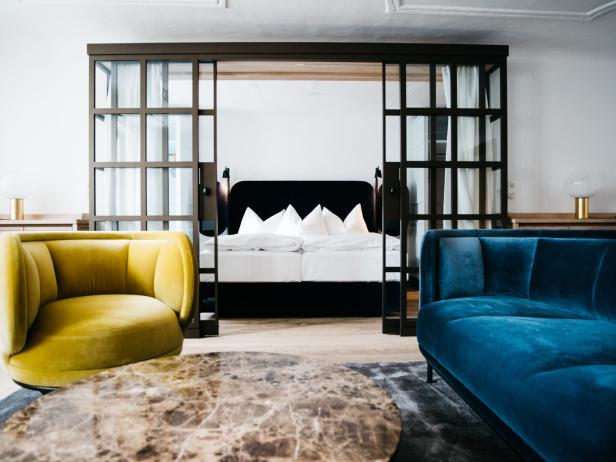 Ein modernes Zimmer mit gelbem Sessel, blauem Sofa, rundem Marmortisch und einem Bett hinter einer Glas-Schiebetür.