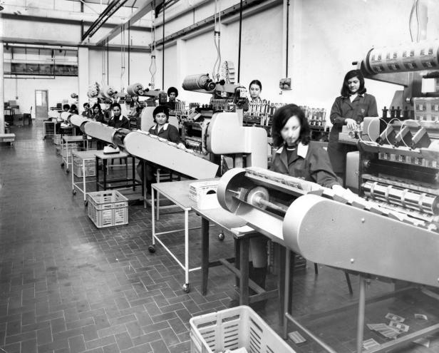 In einer Fabrikhalle arbeiten Frauen an langen Maschinen, möglicherweise in der Zigarettenherstellung.