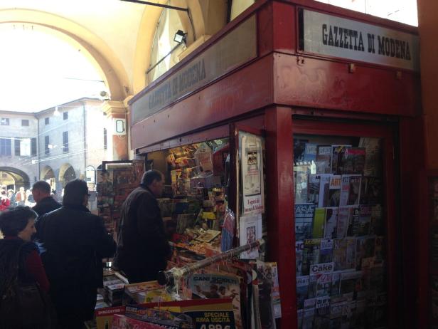 Ein roter Kiosk mit der Aufschrift "Gazzetta di Modena" ist gut gefüllt mit Zeitschriften und Zeitungen.