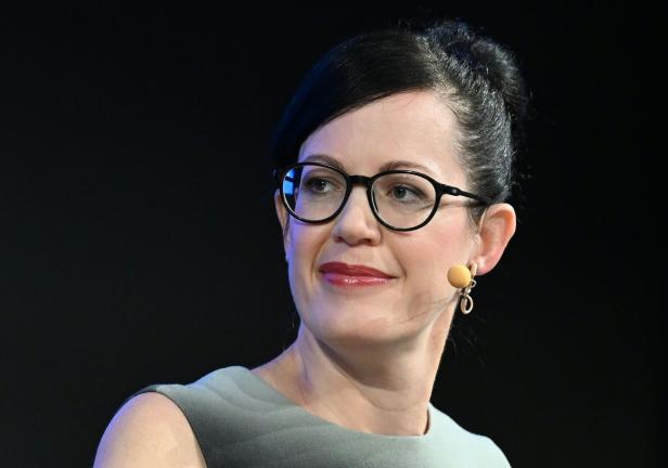 Frau mit dunklen Haaren, Brille und Mikrofon am Ohr blickt zur Seite, trägt ein graues Oberteil.