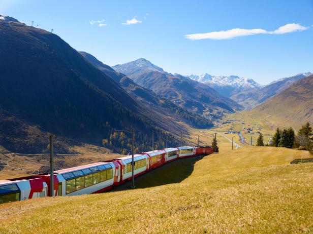 Panoramazug fährt durch ein sonniges Alptal mit schneebedeckten Bergen im Hintergrund.