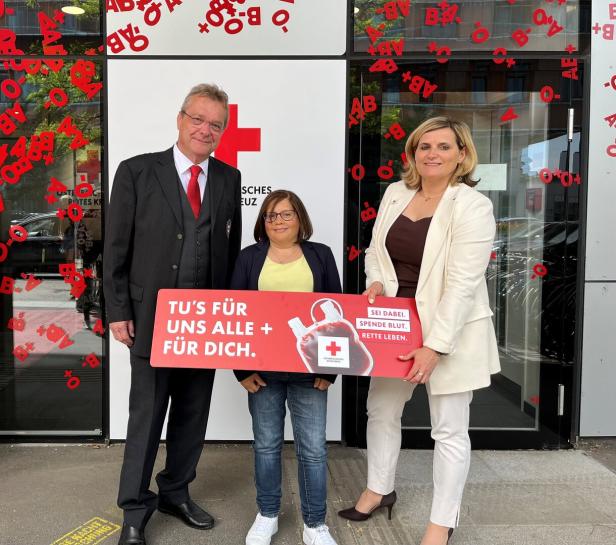 Drei Personen stehen vor einem Gebäude des Deutschen Roten Kreuzes und halten ein Schild zur Blutspendeaktion.