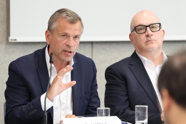 Zwei Männer im Anzug sitzen an einem Tisch, einer spricht gestikulierend ins Mikrofon, der andere hört zu.
