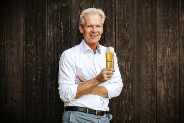 Ein älterer Mann mit weißem Hemd hält ein Glas Bier vor einer dunklen Holzwand und lächelt.