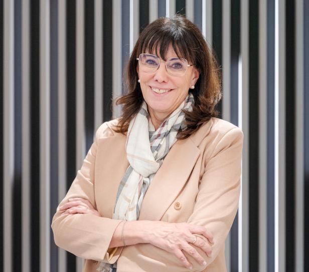 Frau mit braunen Haaren, Brille und beigem Blazer steht lächelnd mit verschränkten Armen vor einem gestreiften Hintergrund.
