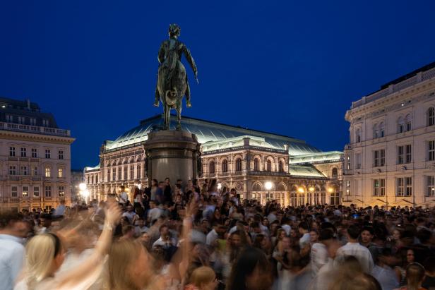 Menschenmenge vor der beleuchteten Wiener Staatsoper bei Nacht, Statue im Vordergrund.