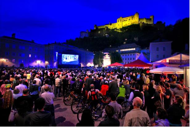 Eine Menschenmenge steht vor einer Bühne, im Hintergrund die Festung Hohensalzburg.