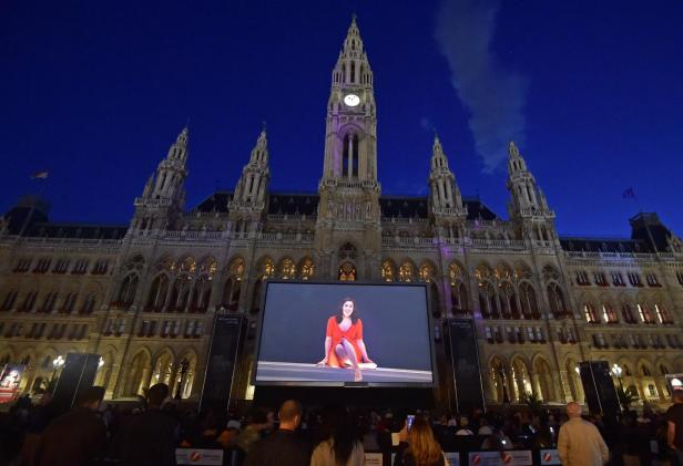 Mehrere Menschen sitzen vor dem Wiener Rathaus und schauen auf eine großflächige Leinwand.
