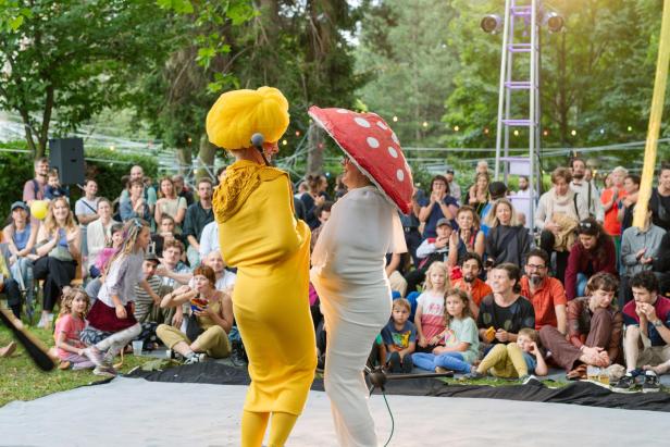 Zwei Künstlerinnen in bunten Kostümen treten vor einem lachenden Open-Air-Publikum im Park auf.