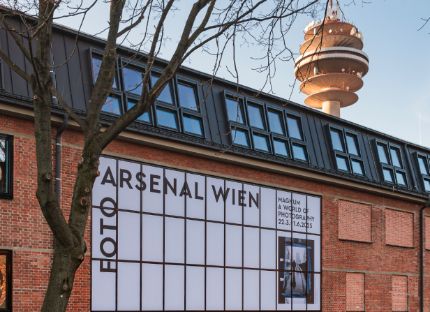 Backsteinfassade des Foto Arsenal Wien mit Ausstellungsplakat, im Hintergrund der Wiener Donauturm.