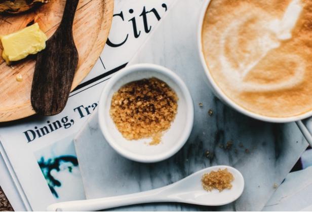 Frühstück mit Cappuccino, braunem Zucker, Butter und Brötchen auf Zeitungsseiten angerichtet.