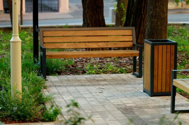 Leere Parkbank aus Holz mit schwarzem Metallgestell neben einem Mülleimer mit Holzverkleidung auf gepflastertem Weg im Freien.