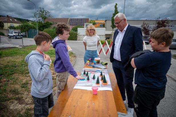 Vier Kinder und ein Erwachsener betrachten gemeinsam ein Bastelprojekt mit kleinen Bäumen und bunten Materialien auf einem Holztisch im Freien.