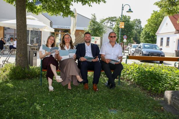 Vier Erwachsene sitzen auf einer Bank im Grünen und halten Broschüren in den Händen, im Hintergrund sind Häuser und Autos zu sehen.