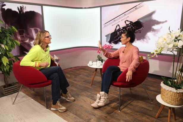 Zwei Frauen sitzen in einem Fernsehstudio in roten Sesseln und führen ein Gespräch vor einem Bildschirm mit der Aufschrift „Herrlich ehrlich“.