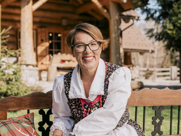 Eine kroatische Gastgeberin in traditioneller Tracht sitzt lächelnd vor einem rustikalen Haus und verkörpert die Initiative „Local Host“.