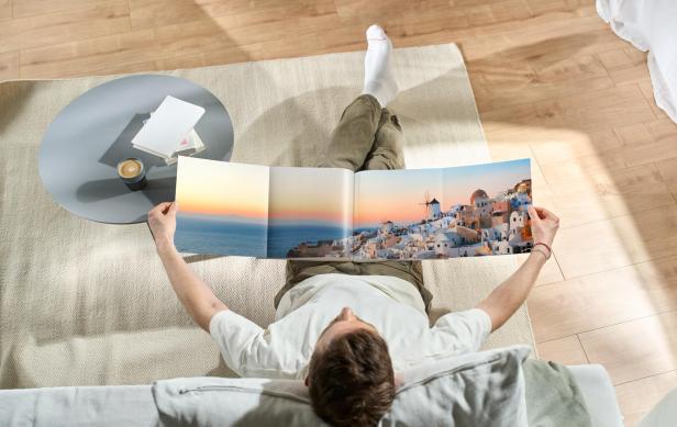 Person sitzt auf einem Sessel und hält ein aufgeschlagenes Fotobuch mit Panoramaaufnahme einer Küstenstadt