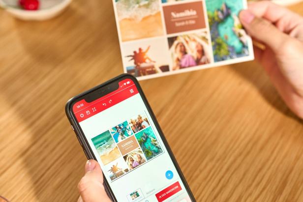 Hände halten eine Postkarte und ein Smartphone mit einer digitalen Ansicht derselben Postkarte auf einem Holztisch