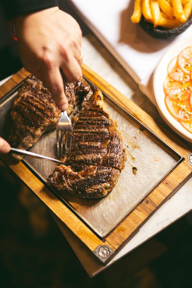 Eine Person schneidet ein gegrilltes Steak auf einem Holzbrett, begleitet von Pommes frites und Tomatensalat.