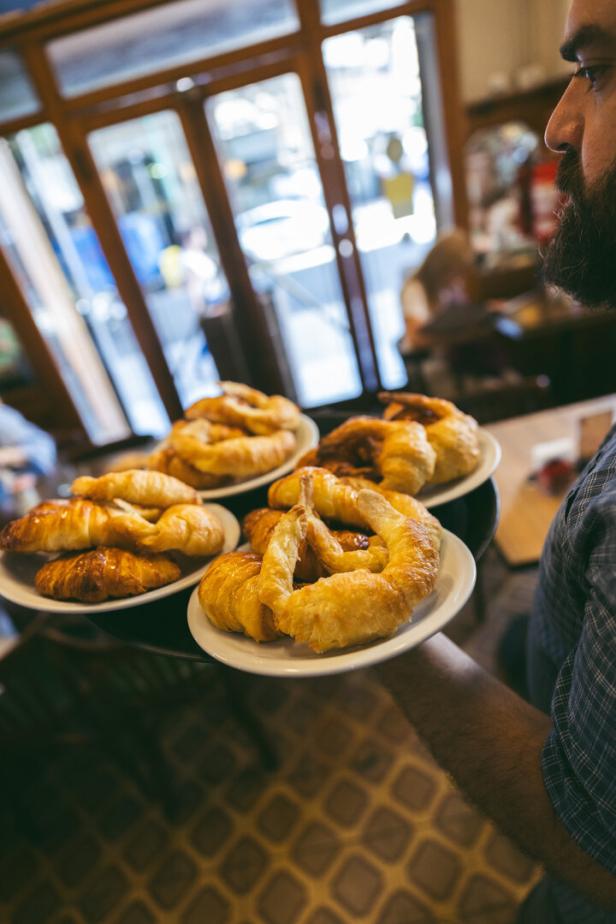 Ein Kellner mit Bart trägt ein Tablett mit mehreren Tellern voller frisch gebackener Croissants durch ein Café.