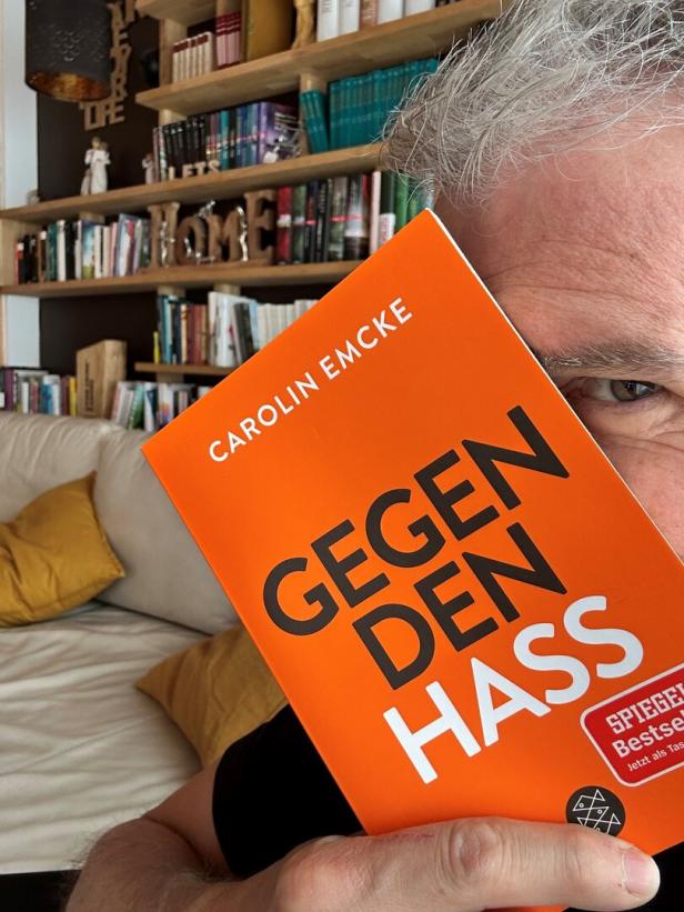 Thomas Raab hält das orange Buch „Gegen den Hass“ von Carolin Emcke vor ihr Gesicht, im Hintergrund ein Bücherregal.