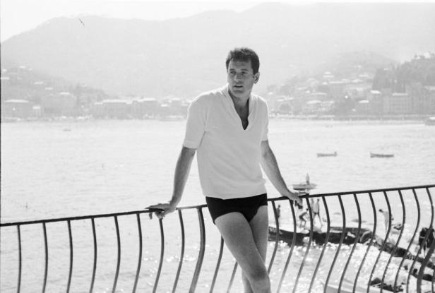 Rock Hudson steht in Badehose und weißem Poloshirt an einem Geländer vor einer Küstenlandschaft.