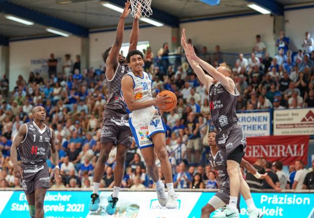 Ein Basketballspieler im weißen Trikot springt zum Korb, während mehrere Gegenspieler in grauen Trikots ihn verteidigen.