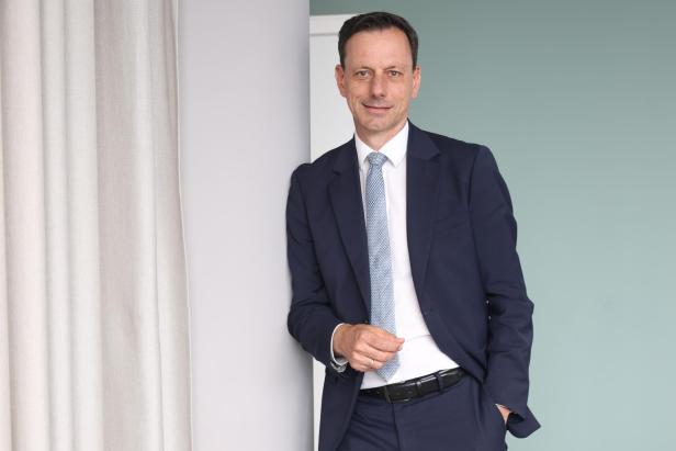 Ein Mann in einem dunkelblauen Anzug mit weißem Hemd und Krawatte lehnt lächelnd an einer Wand.