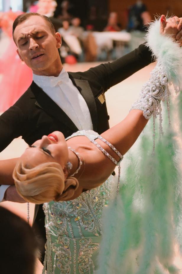 Ein elegant gekleidetes Tanzpaar tanzt eng umschlungen, die Frau trägt ein glitzerndes, mintgrünes Kleid mit Federn.