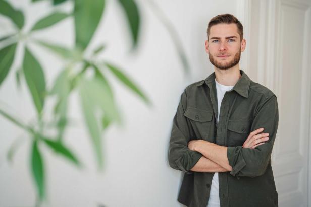 Ein junger Mann mit Bart steht mit verschränkten Armen vor einer weißen Wand, im Vordergrund unscharfe grüne Blätter.