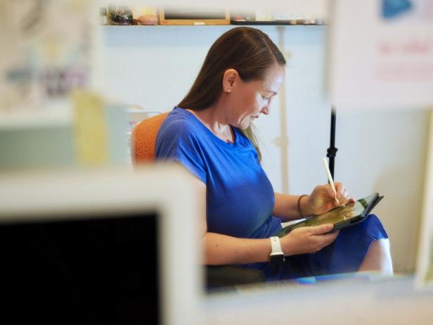 Eine Frau in blauem Kleid sitzt auf einem Stuhl und zeichnet mit einem Stift auf einem Tablet.