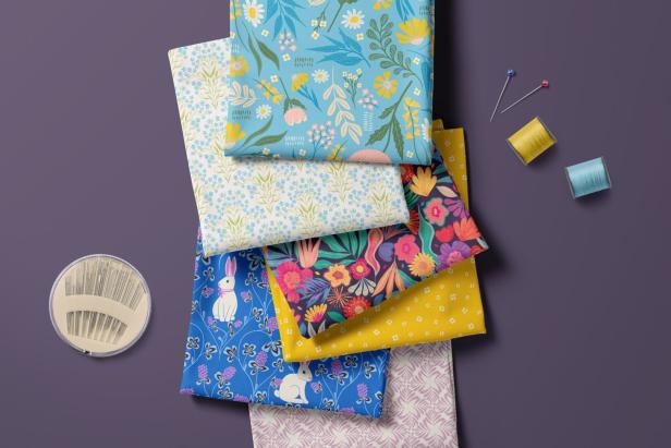Mehrere bunte Stoffstücke mit floralen und tierischen Mustern, Nähnadeln, Garnrollen und Stecknadeln liegen nebeneinander.