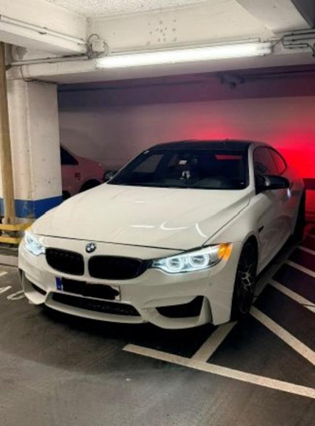 Ein weißer BMW mit eingeschaltetem Licht steht in einem Parkhaus auf einem markierten Parkplatz.