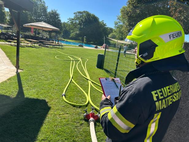 Ein Feuerwehrmann mit gelbem Helm steht auf einer Wiese und überprüft eine Liste neben ausgerollten Schläuchen.