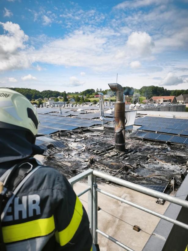 Ein Feuerwehrmann blickt auf ein abgebranntes Dach mit zerstörten Solarpanels und rauchendem Schornstein.