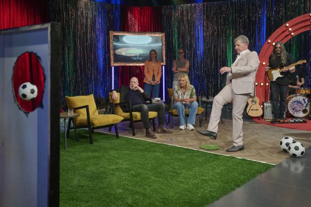 Ein Mann im Anzug schießt einen Fußball durch ein Loch in einer TV-Studioszene.