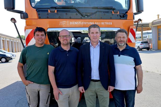 Vier Männer stehen nebeneinander vor einem orangefarbenen Lkw mit der Aufschrift „Dienstleistung.Bau.Betrieb“.