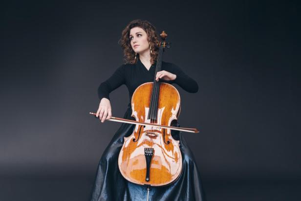 Eine Frau mit lockigen Haaren spielt ein Cello und trägt ein langes schwarzes Kleid vor dunklem Hintergrund.