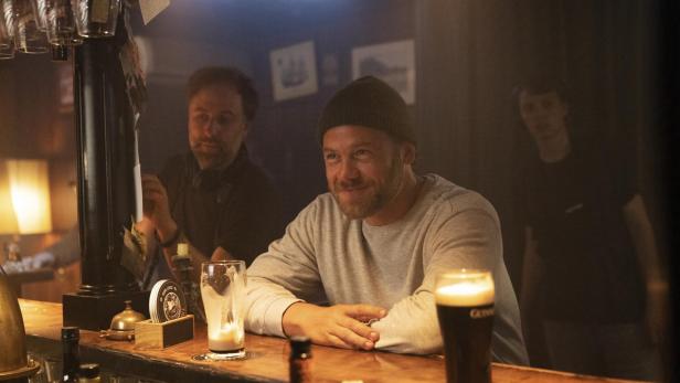 Ein Mann mit Mütze sitzt lächelnd an einer Theke, vor ihm stehen zwei Gläser Bier, im Hintergrund zwei weitere Personen.