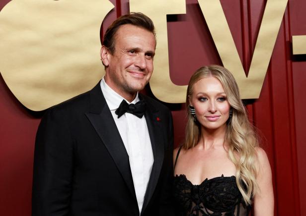 Jason Segel und Kayla Radomski posieren elegant vor einem roten Hintergrund mit goldenen Schriftzeichen.