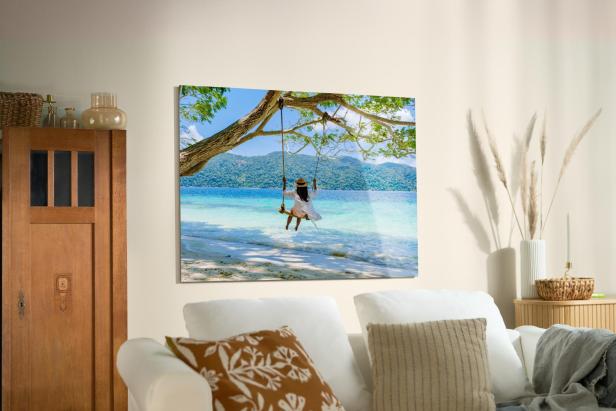 Ein großformatiges Wandbild mit einem Urlaubsfoto von einer Frau auf einer Schaukel am Strand bringt sommerliche Erinnerungen in ein gemütlich eingerichtetes Wohnzimmer.