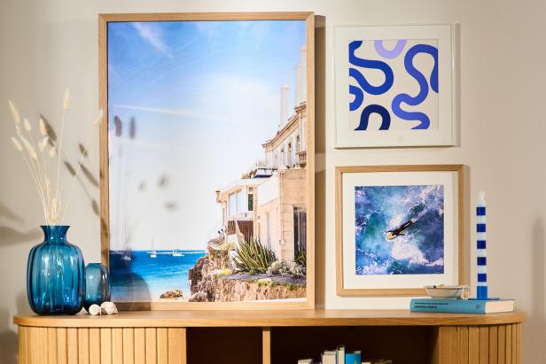 Holzsideboard mit gerundeten Kanten, darauf ein blauer Glasvase, drei gerahmte Bilder an der Wand, darunter ein großes mit Meeresblick, und verschiedene Bücher und Dekorationsobjekte in den offenen Fächern
