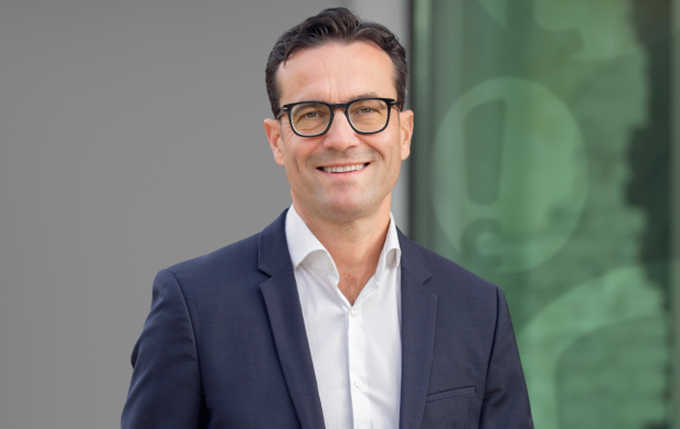 Mann im Anzug mit Brille lächelt vor modernem, grünlichem Hintergrund.