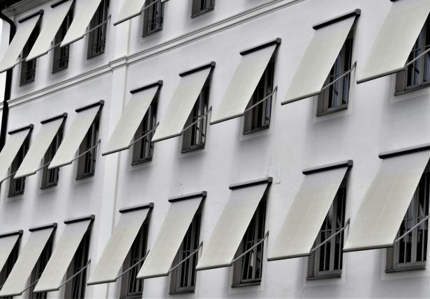 Mehrere Fenster mit schräg gestellten, hellen Markisen an einer weißen Hausfassade.
