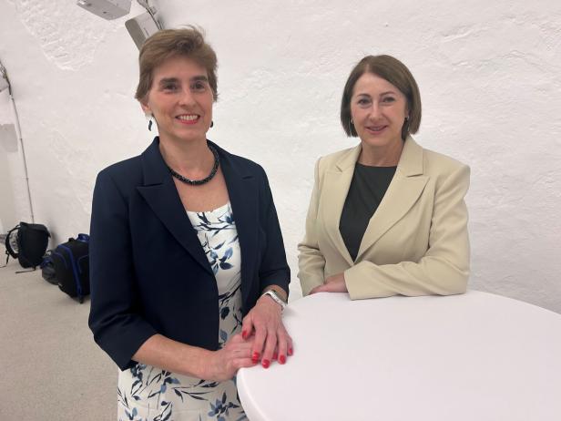 Zwei Frauen stehen an einem runden Tisch vor einer weißen Wand und lächeln in die Kamera.