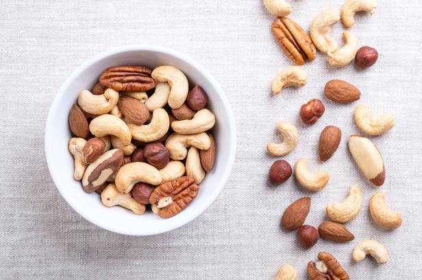Verschiedene Nüsse wie Cashews, Mandeln, Pekannüsse und Haselnüsse liegen in einer weißen Schale und daneben auf einem hellen Stoff.